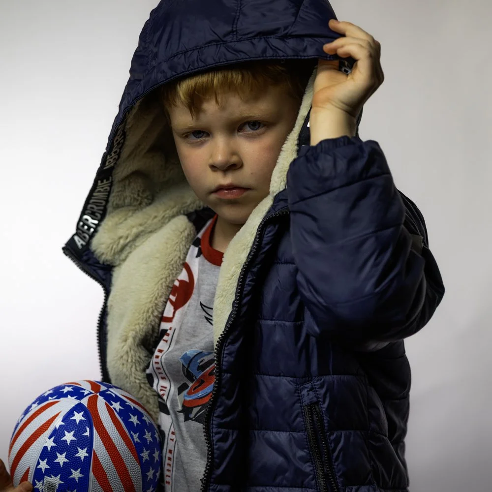 A young boy with light brown hair and blue eyes wearing a navy jacket with a hood, holding a basketball decorated with an American flag design, against a plain gray background.