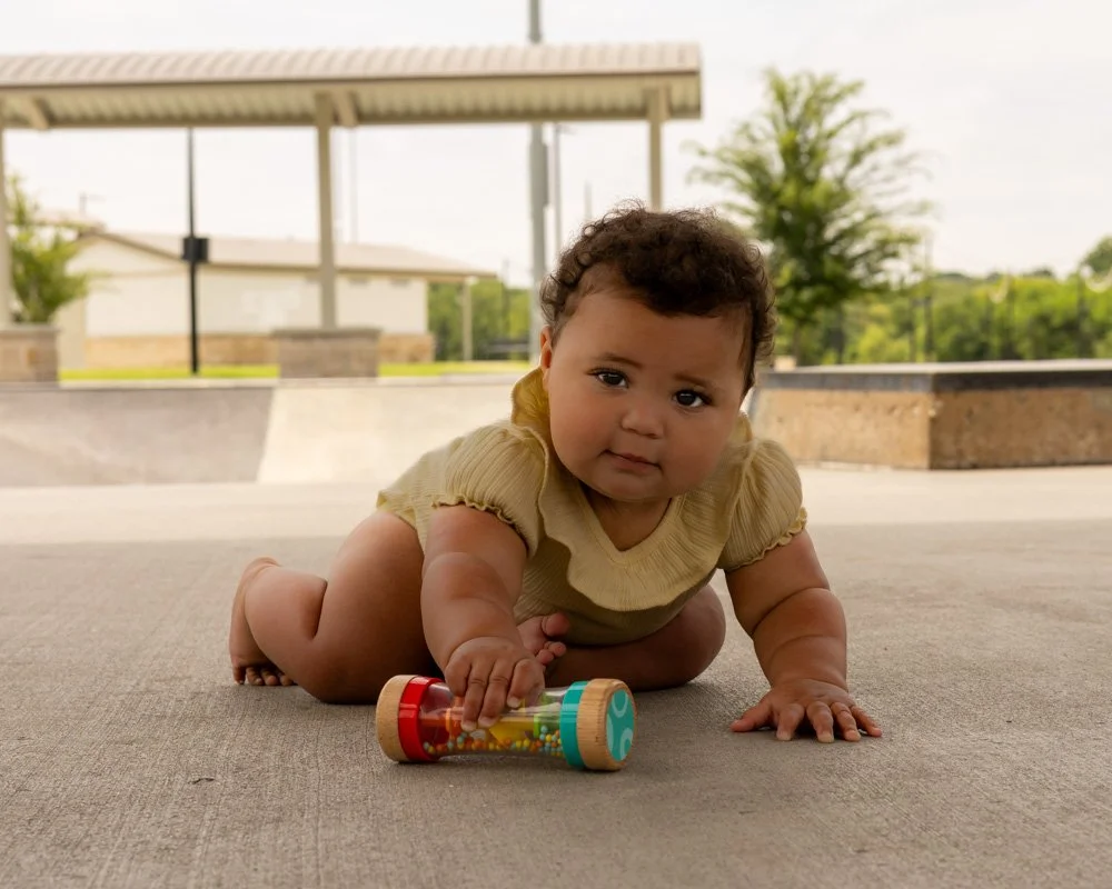 Young child crawling on a concrete surface outdoors, playing with a colorful toy, with trees and a picnic shelter in the background.
