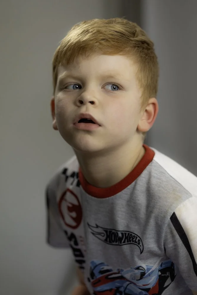 A young boy with red hair and blue eyes looking to his left, wearing a gray Hot Wheels t-shirt with red trim and car graphics.