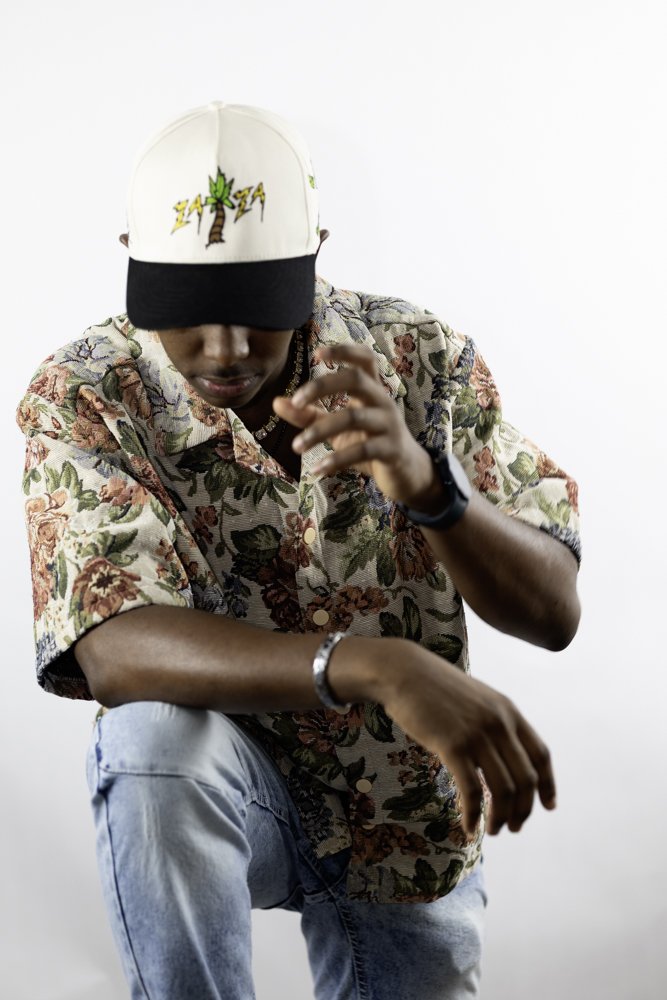 A young man wearing a floral shirt, light blue jeans, and a white baseball cap with a palm tree and 'ZAM ZAM' on it. He is sitting and looking down, adjusting his watch or bracelet against a plain white background.