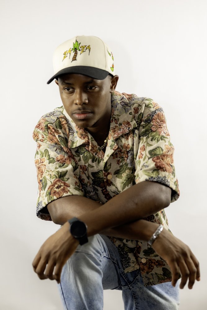 A young man wearing a beige baseball cap with a palm tree and lightning bolt design, a floral patterned shirt, light jeans, a black watch, and a silver bracelet, sitting with arms crossed against a plain white background.