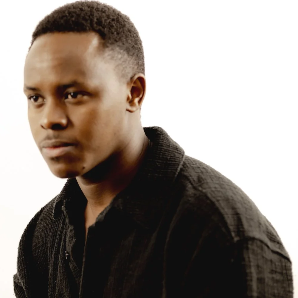Close-up of a young African American man with short curly hair wearing a black textured shirt, looking serious against a white background.