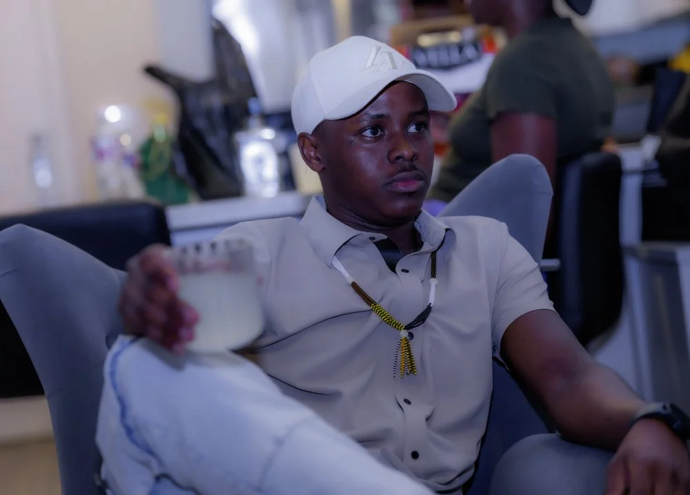 A young man wearing a white baseball cap and a beige shirt, holding a glass of milk, sitting on a gray couch in a casual indoor setting.