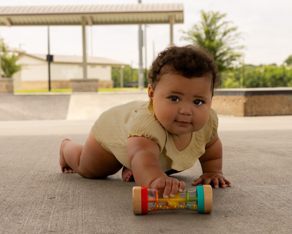 A baby crawling on a concrete surface outdoors, holding a colorful toy with beads and gears, with trees and a shelter in the background.