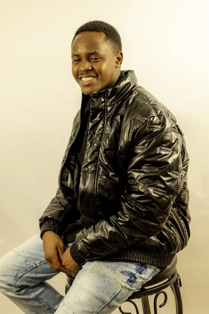 Young man smiling, wearing a shiny black bomber jacket, light-wash jeans, sitting on a black metal stool against a plain off-white background.