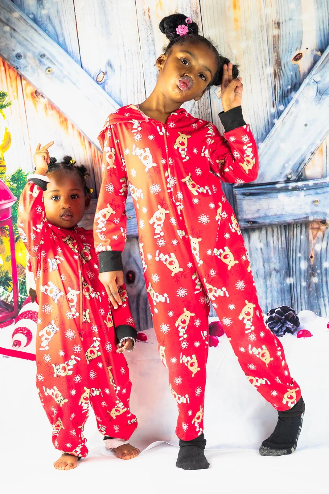 Two young girls in matching red Christmas-themed pajamas with reindeer and snowflake patterns, posing in front of a winter holiday backdrop with decorated Christmas tree and snow.