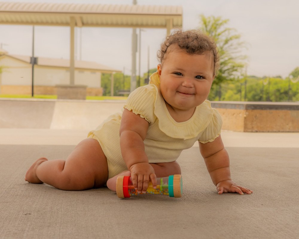 A smiling baby crawling outdoors on a flat surface, holding a colorful toy roller, near a park or playground with trees and structures in the background.