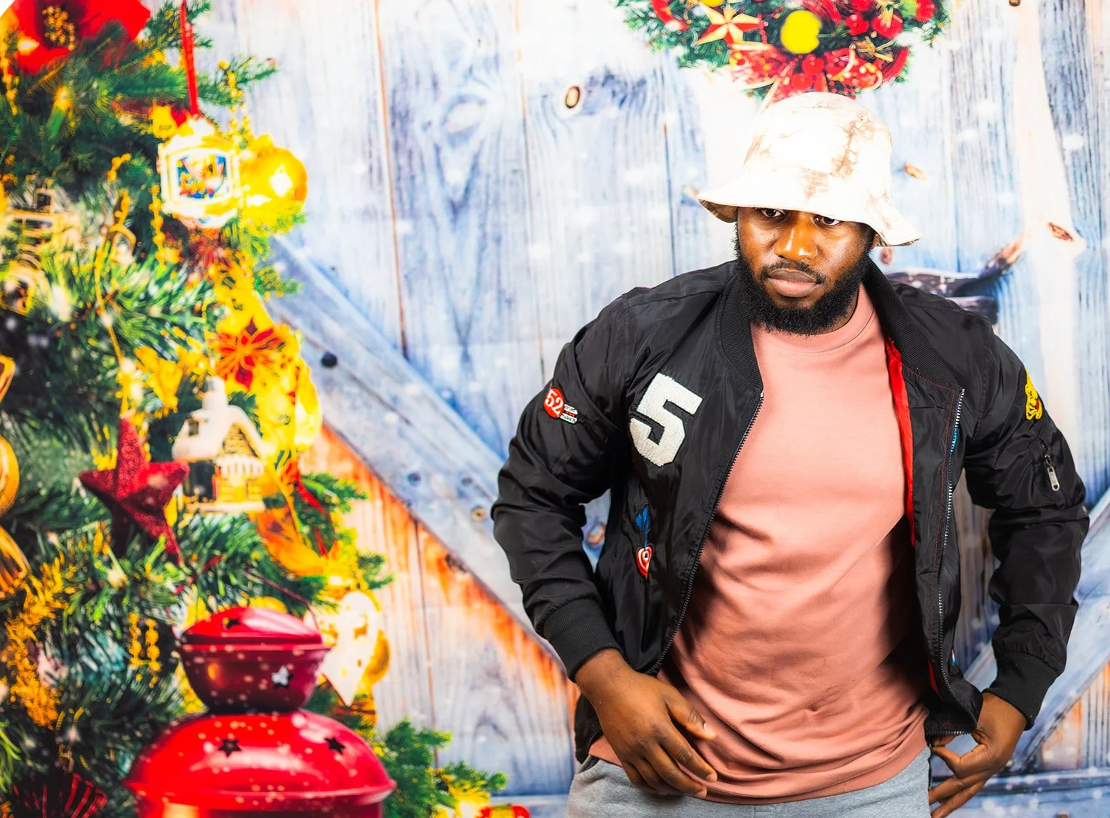 A man wearing a white bucket hat, pink T-shirt, and black jacket stands next to a decorated Christmas tree with ornaments and lights in a festive room.