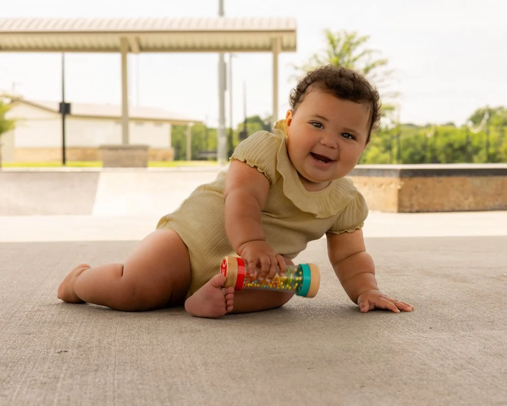 A happy baby lying on a concrete surface outdoors, holding a colorful rattle, with a park and trees in the background.