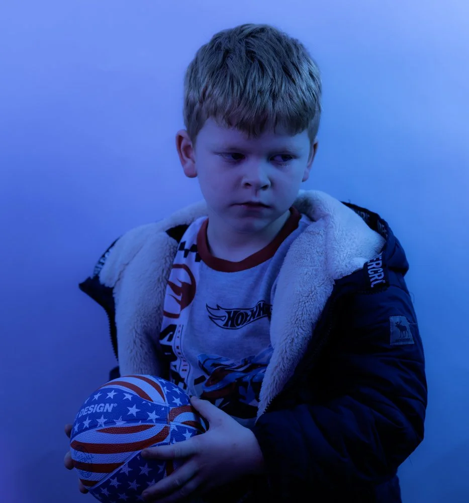 A young boy with red hair wearing a jacket with a furry lining, holding a ball with a patriotic design of red, white, blue, and stars, looking off to the side with a serious expression against a blue background.