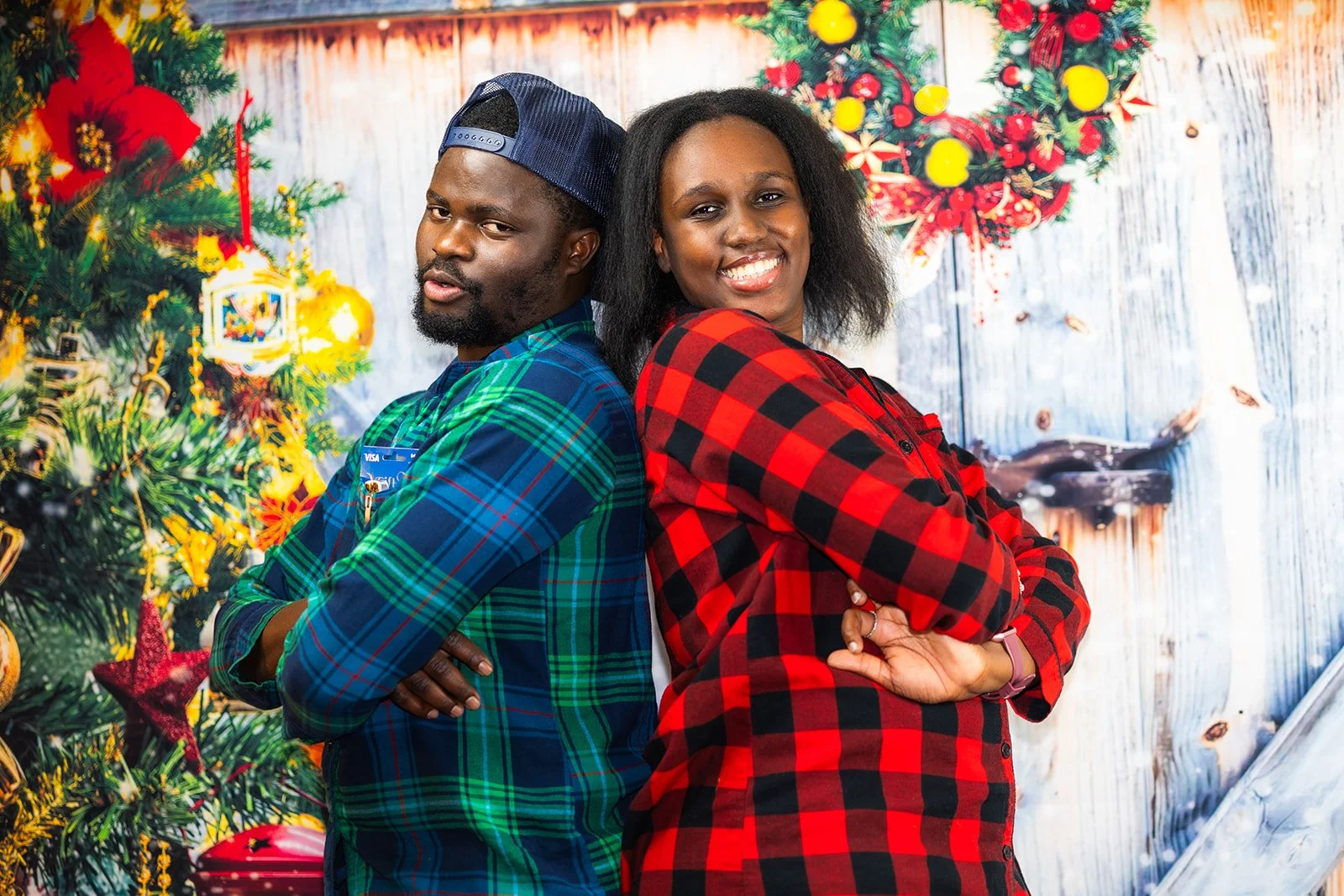 A man and woman standing back-to-back with arms crossed, smiling, in front of Christmas decorations, including a Christmas tree with ornaments and a wreath on a wooden wall.