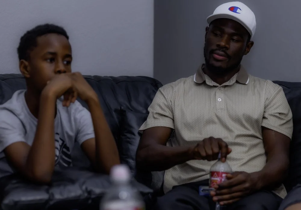An adult man and a young boy sitting on a black sofa in a room with gray walls. The man is wearing a white baseball cap and a beige polo shirt, holding a water bottle, and looking at the boy. The boy is wearing a white t-shirt and has a thoughtful ex