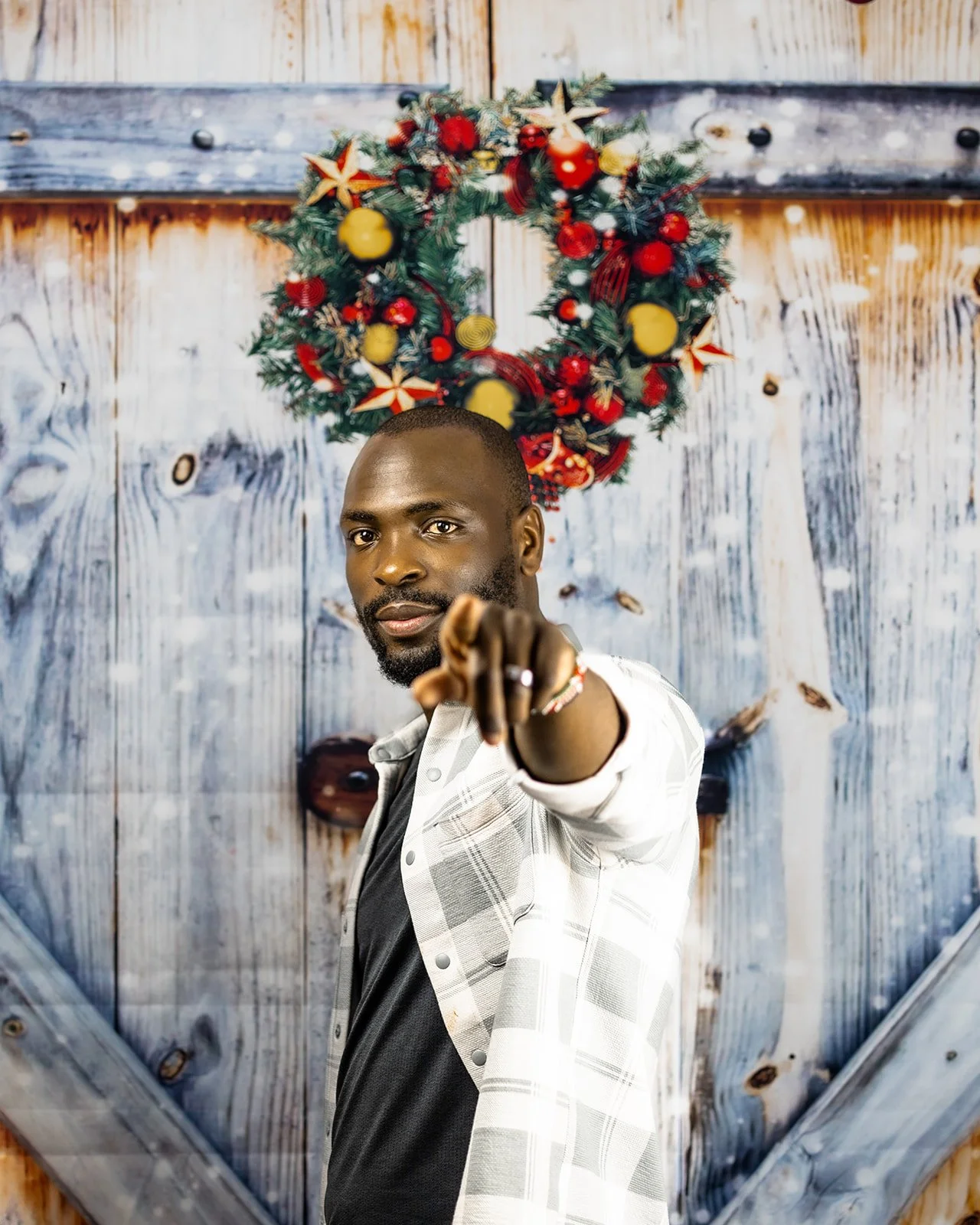 A man points at the camera in front of a wooden wall with a Christmas wreath hanging above his head.