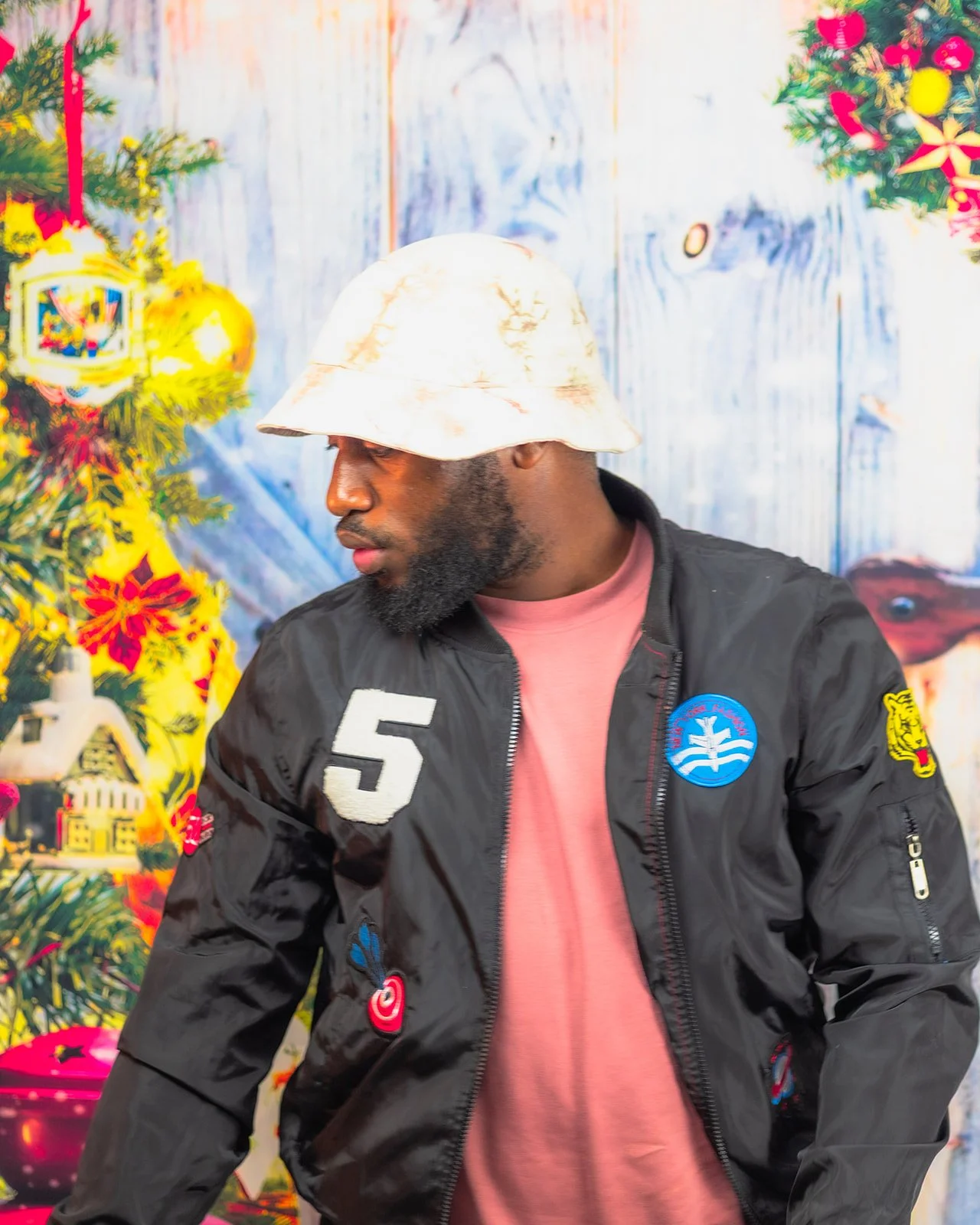 A man wearing a beige bucket hat, pink t-shirt, and a black bomber jacket with patches, standing in front of a decorated Christmas tree and colorful holiday background.