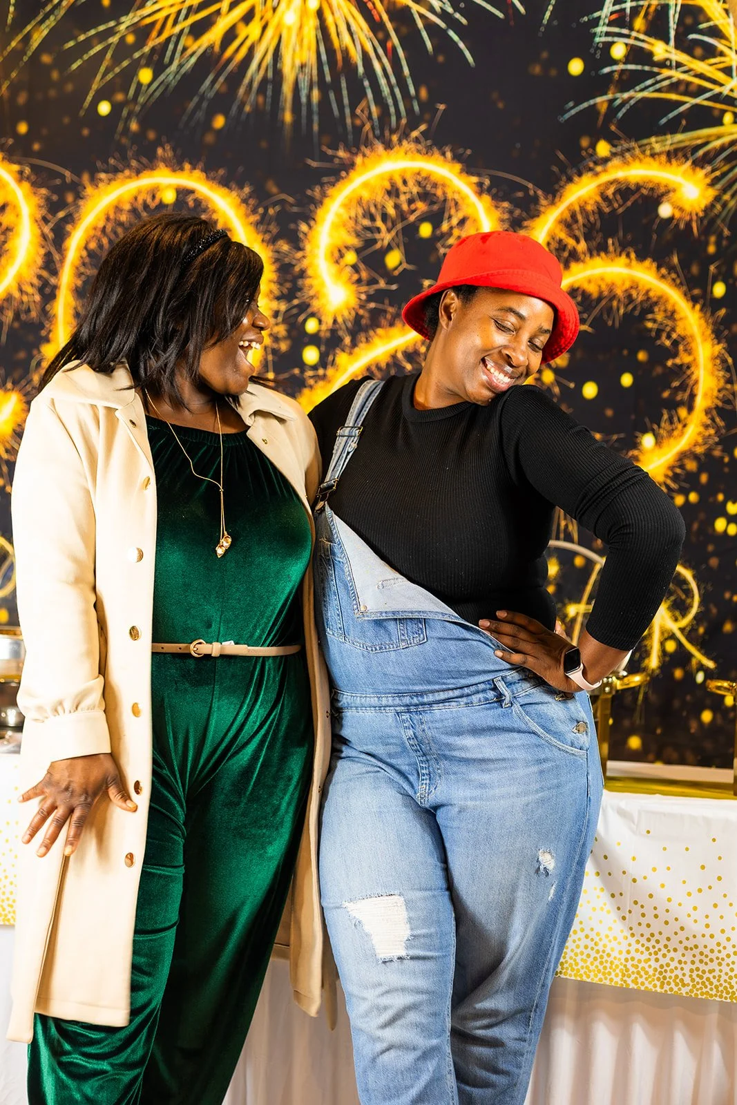 Two women celebrating at a party with fireworks in the background. One woman is dressed in a green velvet jumpsuit with a cream-colored coat, and the other is wearing a red hat, black top, and ripped jeans. They are smiling and enjoying the moment.