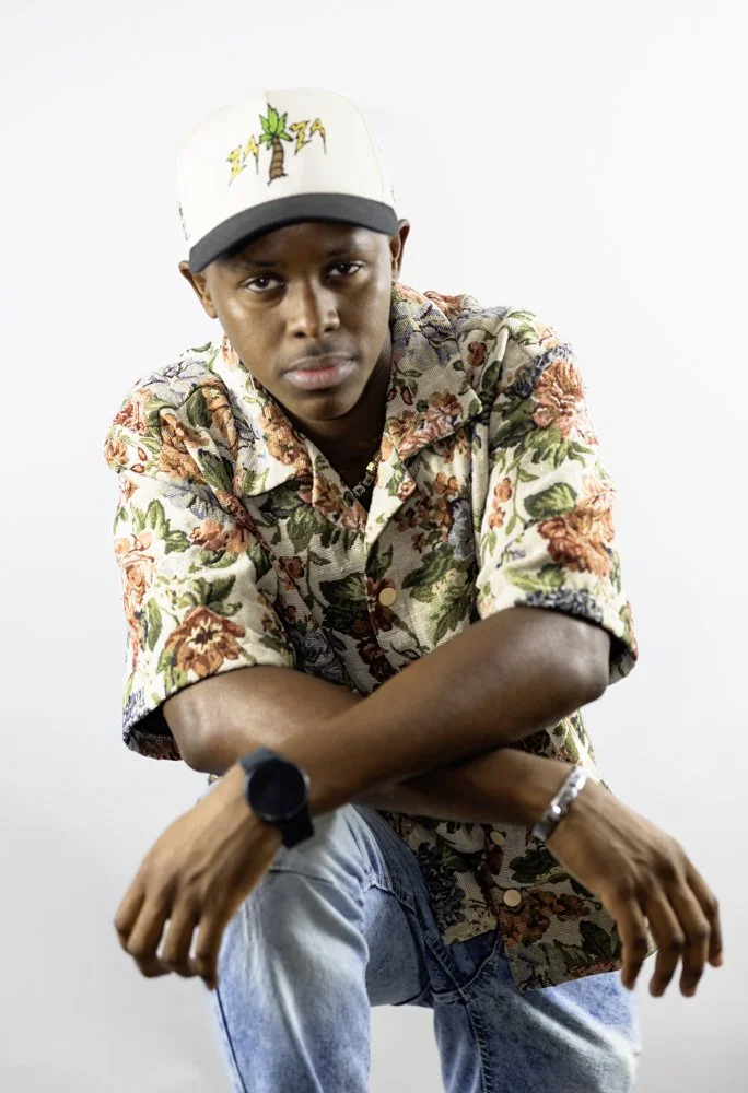 A young man wearing a floral button-up shirt, a white cap with a palm tree and lightning bolts, a watch, and a bracelet, sitting against a plain white background.