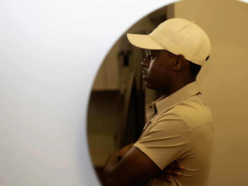 A man wearing a beige baseball cap and beige polo shirt, standing with arms crossed, looking to his left through a round opening in a wall.