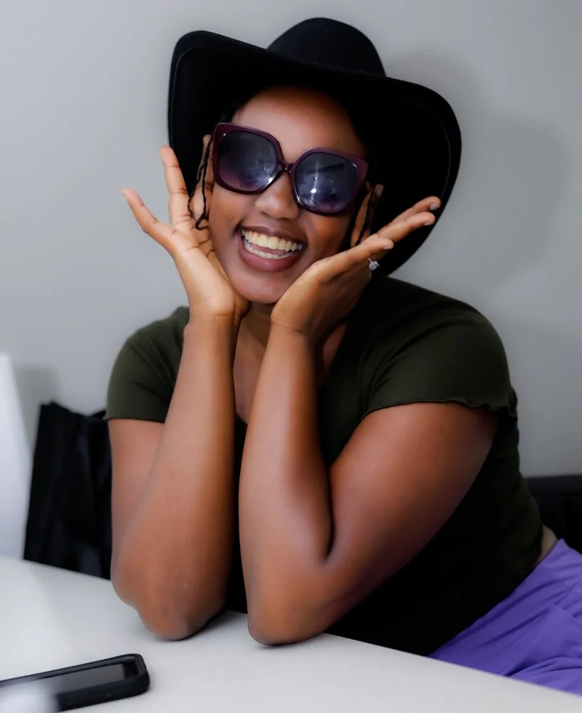 A woman wearing a black hat and large sunglasses, smiling and posing with her hands on her cheeks.