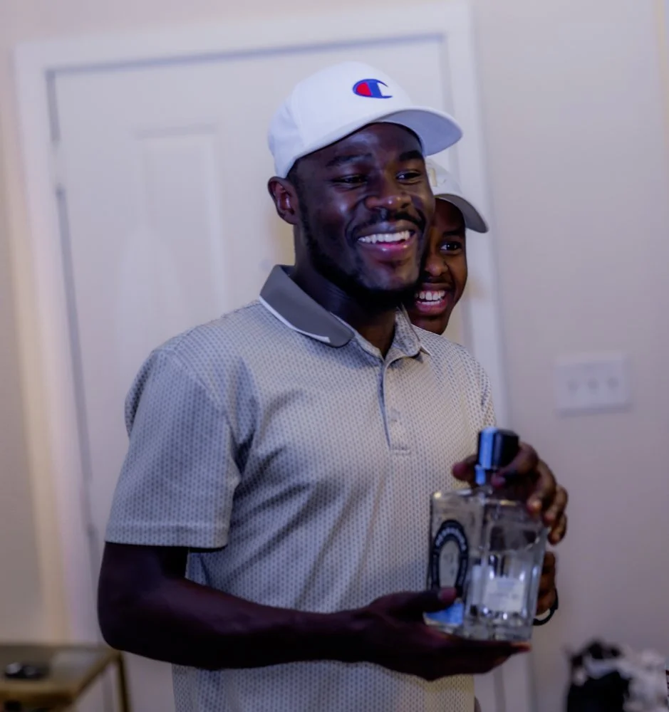 A man wearing a white baseball cap and a gray polo shirt is smiling and holding a bottle of alcohol in his hand. A child is partially visible behind him, also smiling.