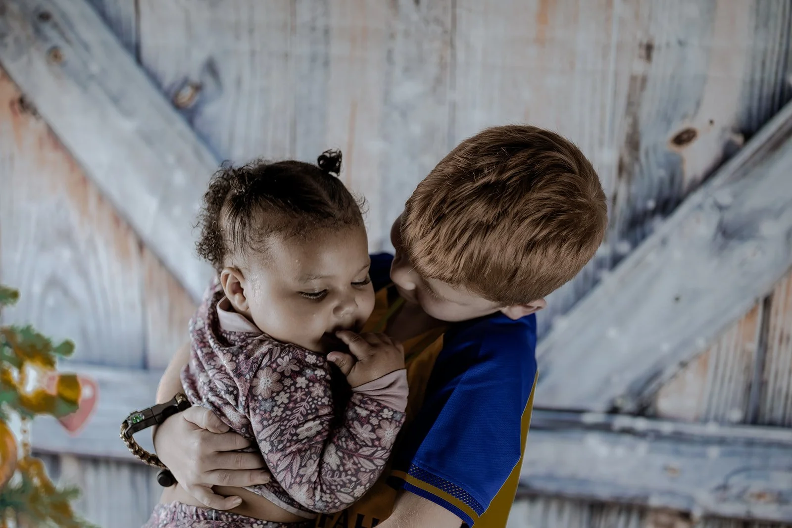A young boy holding a girl with curly hair close to his face, both looking at each other affectionately in an indoor setting with wooden walls in the background.