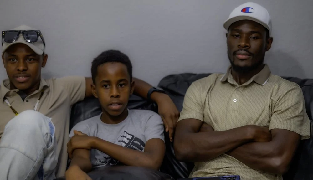Three males sitting on a black leather couch against a light gray wall, with neutral expressions. The male on the left is wearing a beige shirt, sunglasses on his head, and a baseball cap. The boy in the middle has a gray T-shirt and is sitting with 