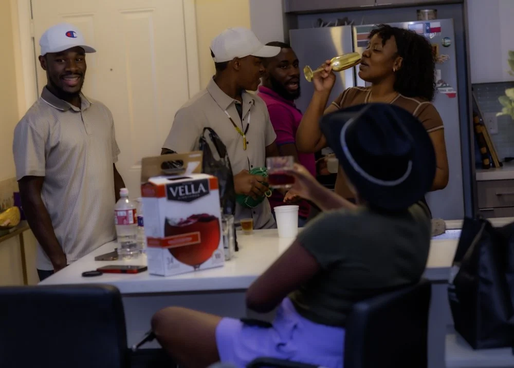 Group of five friends gathered in a kitchen, socializing and drinking beer at a party, with a woman drinking from a beer bottle and a man in a hat holding a drink.