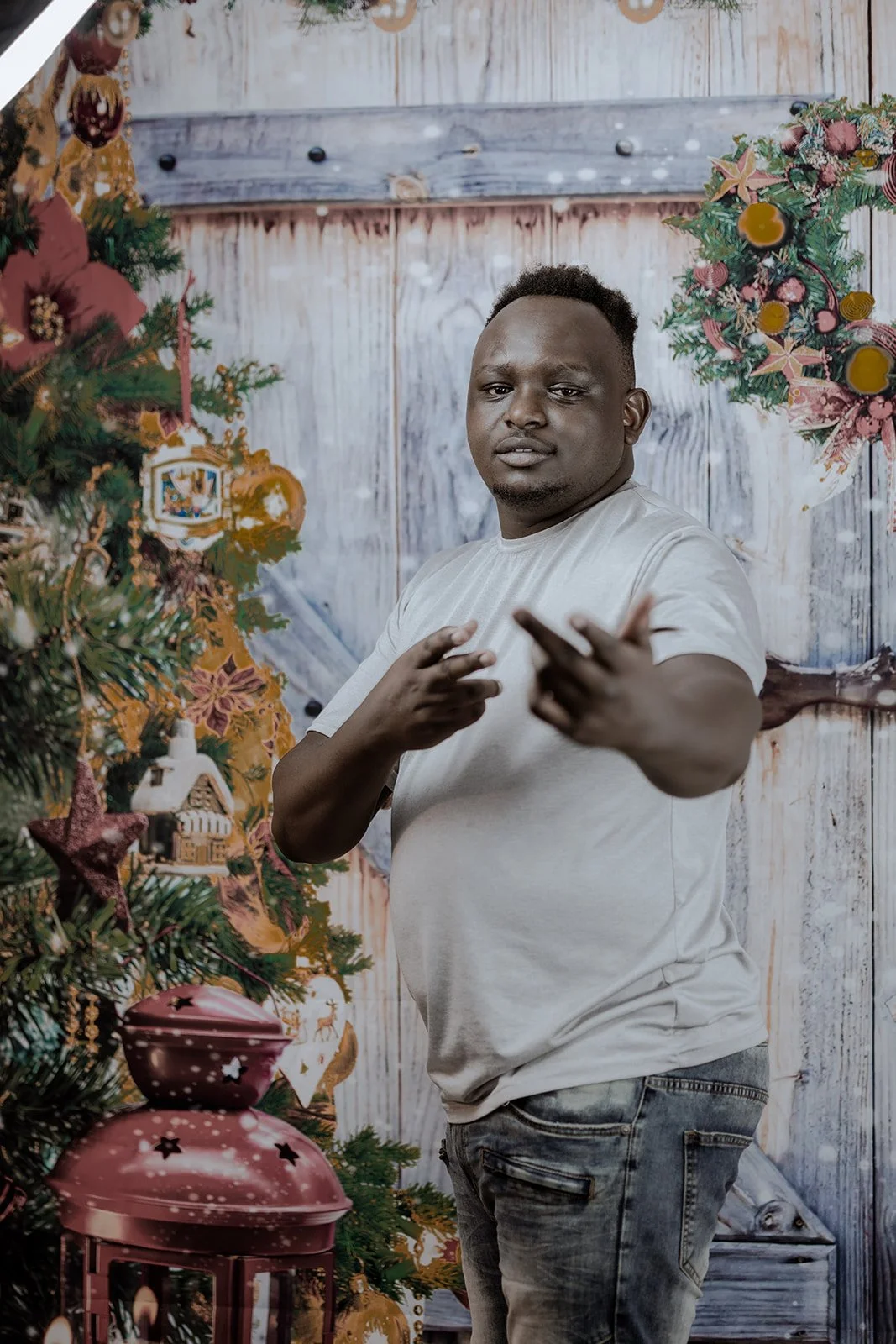 A man standing beside a decorated Christmas tree with ornaments, stars, and a pink lantern, pointing towards the camera with a confident expression, in a rustic, holiday-themed setting.