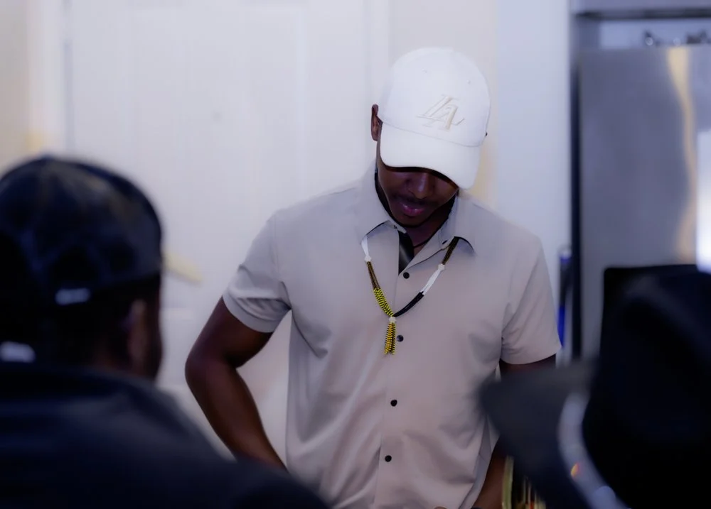 A man wearing a beige cap, beige short-sleeve shirt, and a beaded necklace, standing and speaking to two other men. The background shows a white wall and a refrigerator.