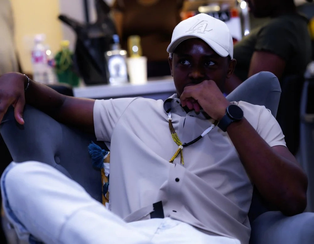 A young man wearing a white cap and beige polo shirt sitting on a couch, resting his arm on the back, with a thoughtful expression, in a casual indoor setting.