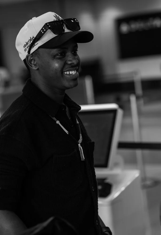 Young man smiling, wearing a baseball cap with sunglasses on top, in an indoor setting with computers in the background.