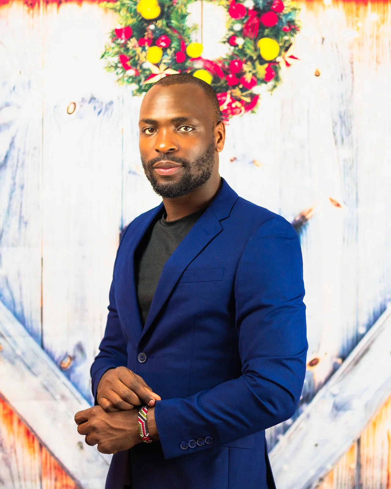 A man in a blue blazer and black shirt standing in front of a decorated wooden background with a wreath of red and yellow ornaments.