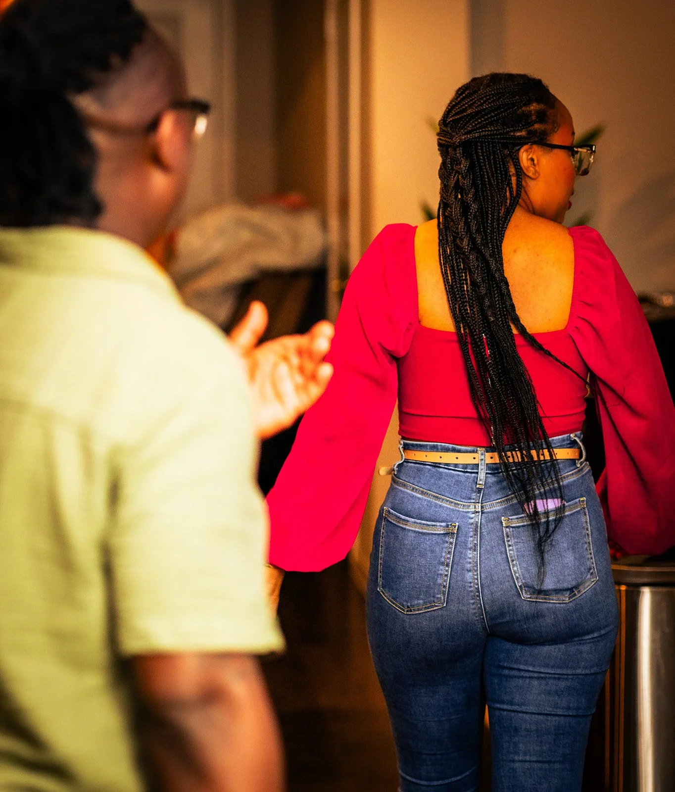 A woman with long braided hair, wearing glasses, a pink top with puffed sleeves, and blue jeans, is standing with her back to the camera. Another person is blurred in the foreground, facing her.