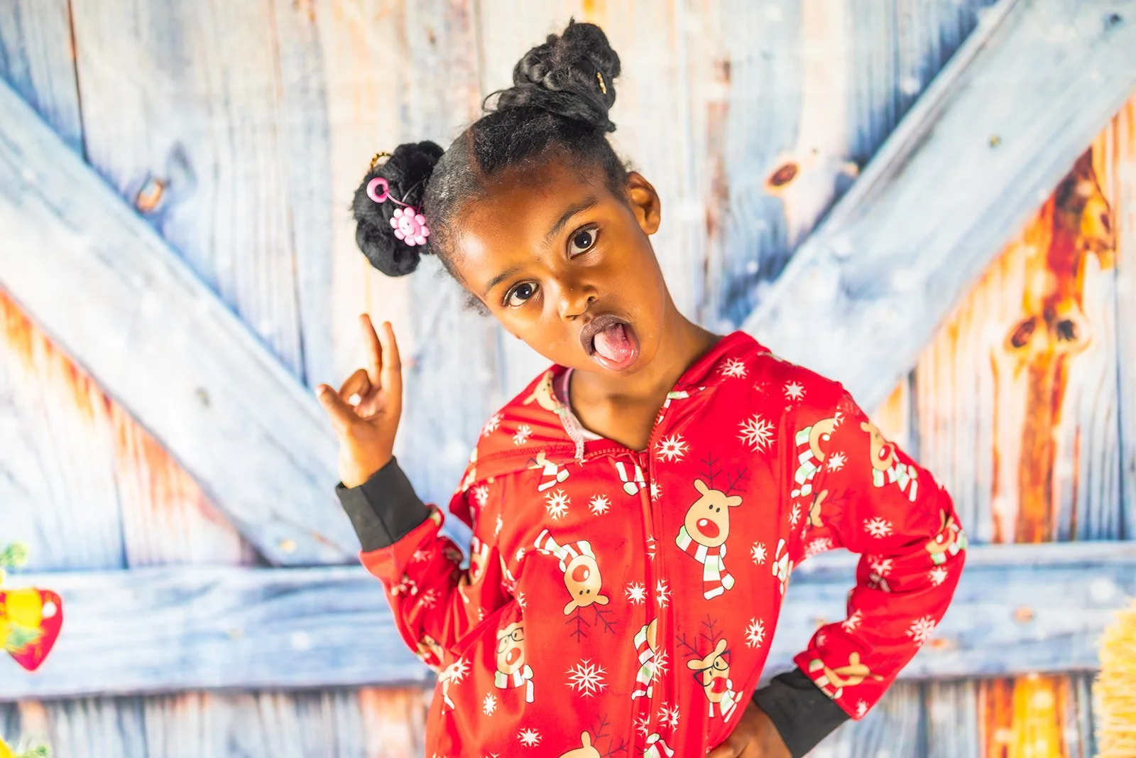 A young girl with braided hair, decorated with pink hair accessories, makes a surprised expression and points at her head. She is wearing a red Christmas-themed jacket with reindeer and snowflakes, standing against a colorful, rustic wooden background.