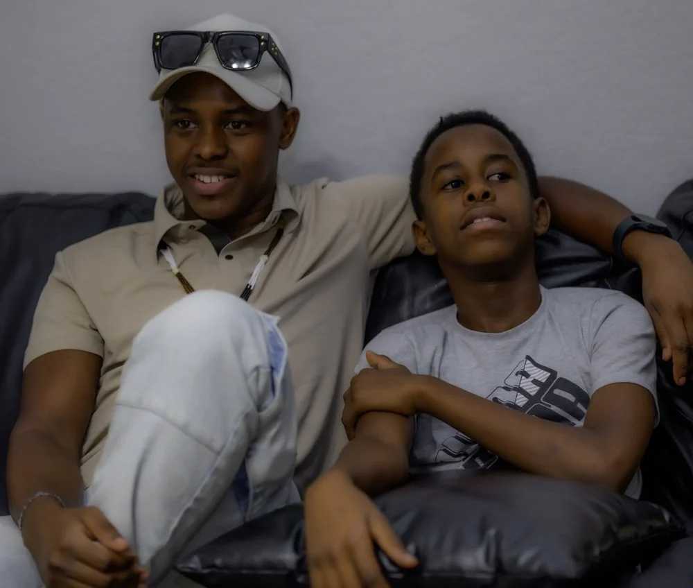 Two young males sitting close together on a black leather couch, one with sunglasses on his head and the other with a neutral expression.