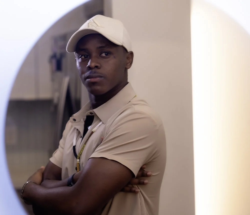 A young man in beige uniform and cap looking into a mirror in a room.
