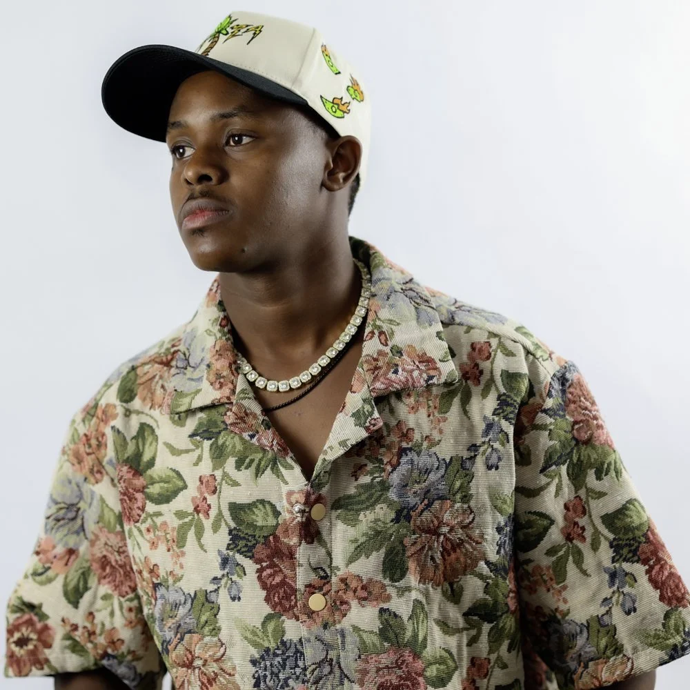 A young man wearing a floral button-up shirt, a pearl necklace, and a baseball cap with colorful designs, looking to the side against a plain white background.