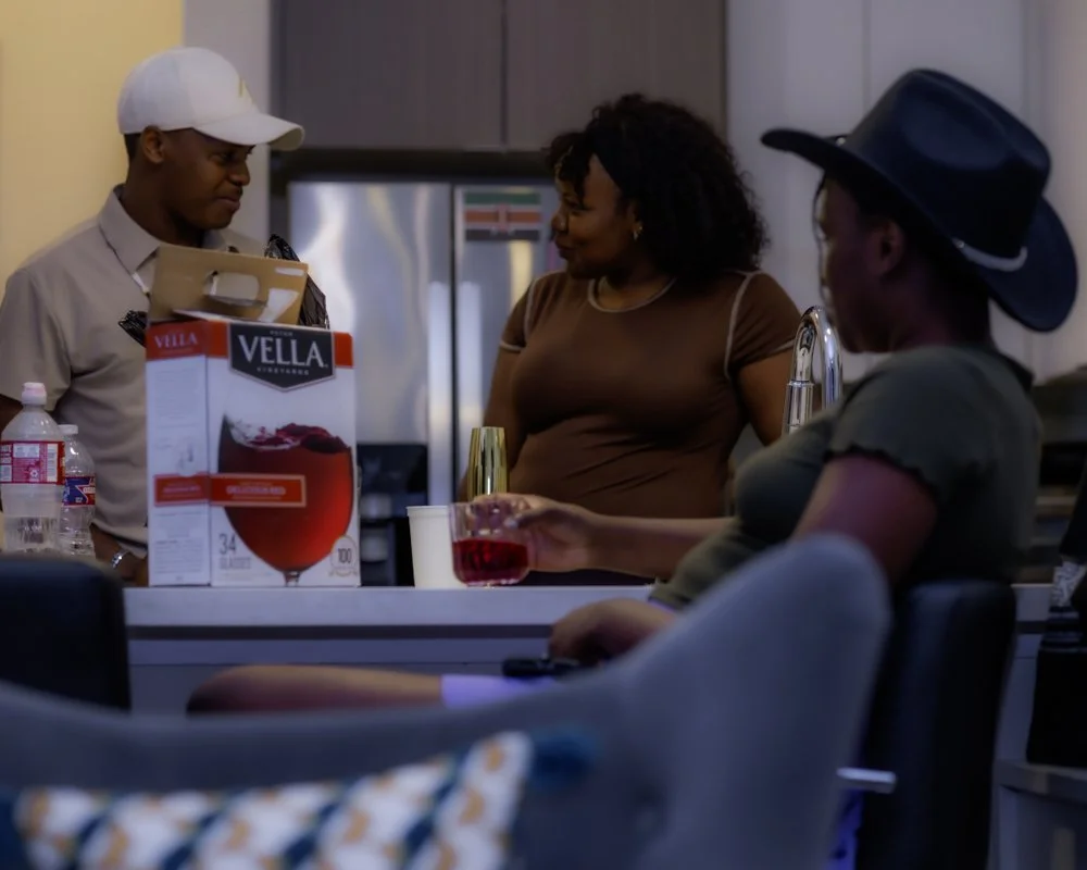 A woman sitting at a kitchen counter holding a glass of red wine, while a man in a white cap and a woman with curly hair and a black hat stand nearby, engaging in conversation.