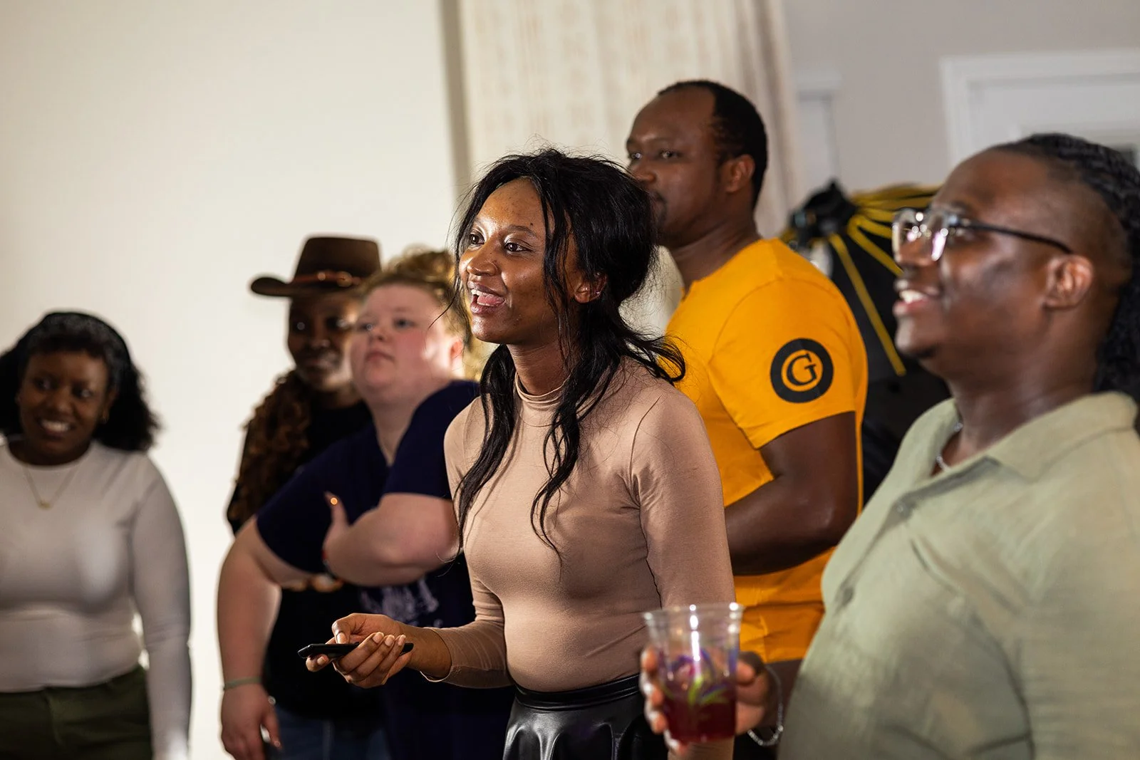 Group of diverse people standing and talking indoors, some holding drinks, engaged in conversation.