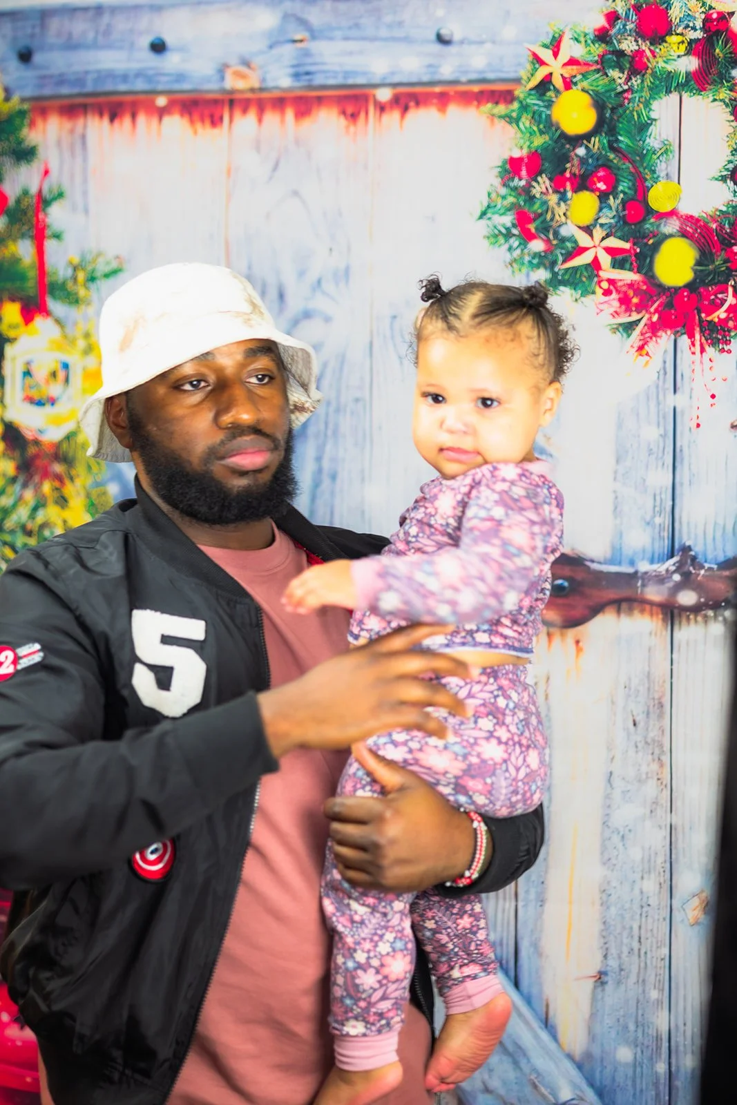 A man with a beard wearing a white bucket hat and a black jacket holding a young girl in colorful pajamas with a floral pattern. The background features Christmas decorations, including a wreath and festive ornaments.