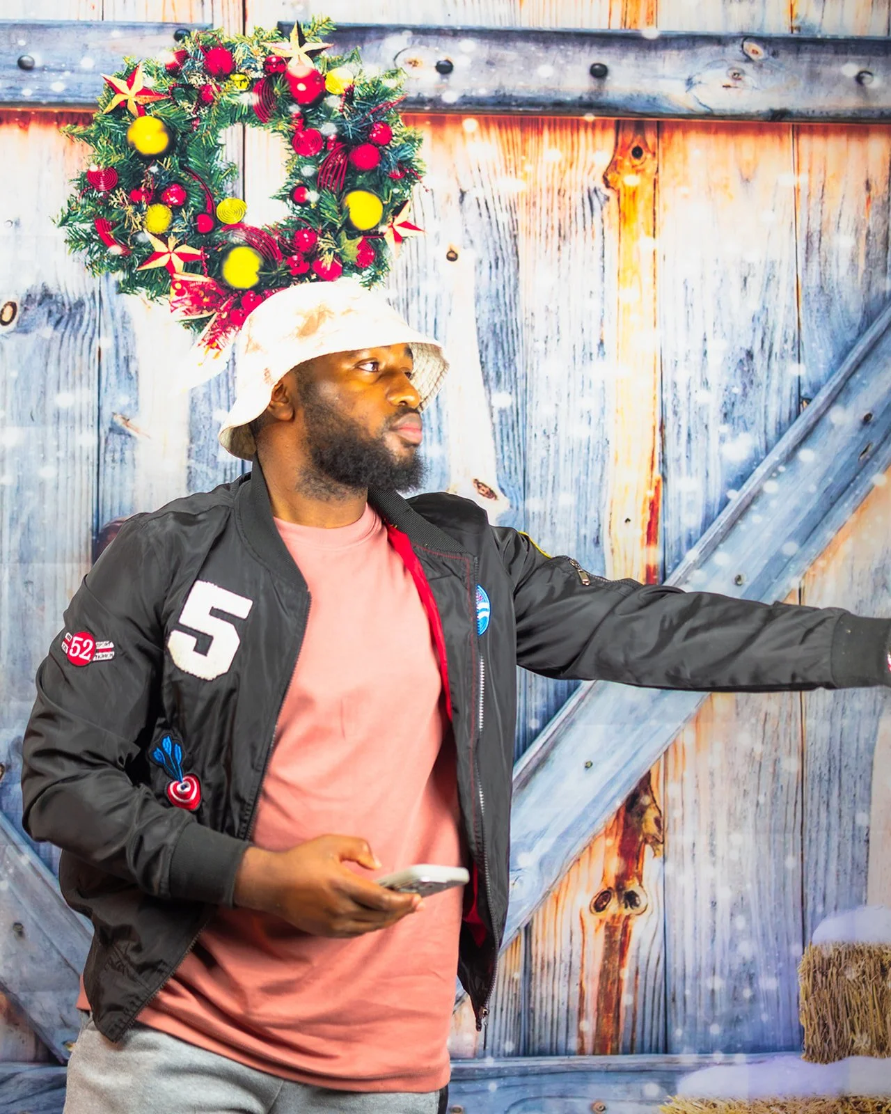 A man wearing a black bomber jacket with patches, a pink T-shirt, and gray sweatpants, standing in front of a wooden wall with a Christmas wreath on a hat on his head, holding a phone in his left hand, with snow falling around him.
