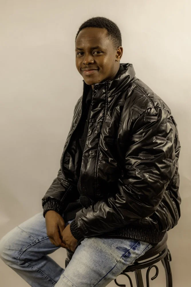 Young man sitting on a stool, wearing a shiny black bomber jacket and light blue jeans, smiling softly at the camera, against a plain white background.