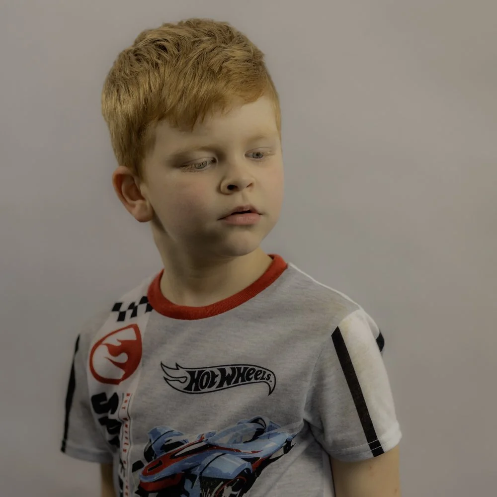 A young boy with red hair wearing a Hot Wheels themed t-shirt, looking to the side with a neutral expression in front of a plain, light-colored wall.