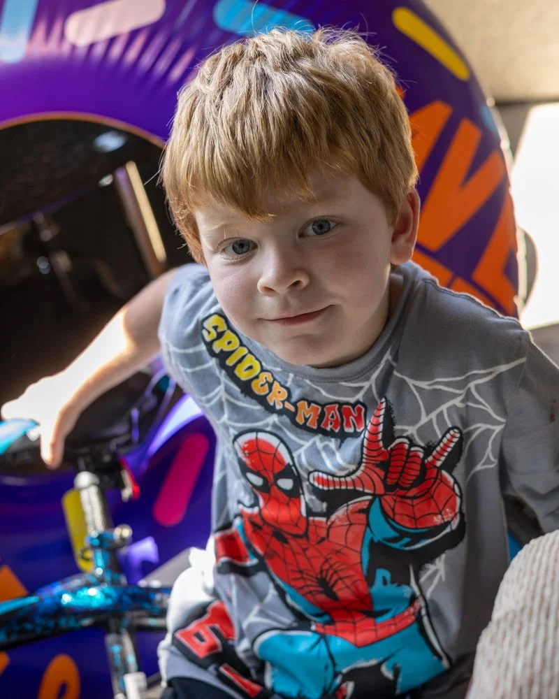 A young boy with red hair and blue eyes sitting on a purple and orange ride or play equipment, wearing a Spider-Man t-shirt.