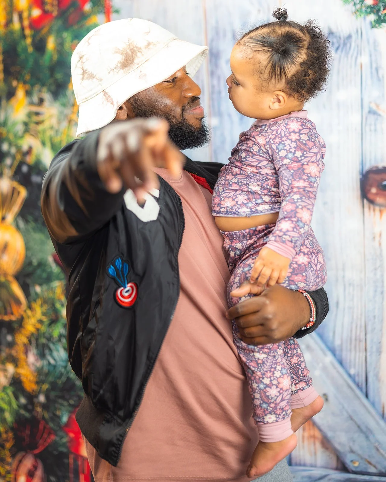 A man holding a young girl in his arms, pointing at something, standing in front of a colorful background with plants and a blue wooden wall.