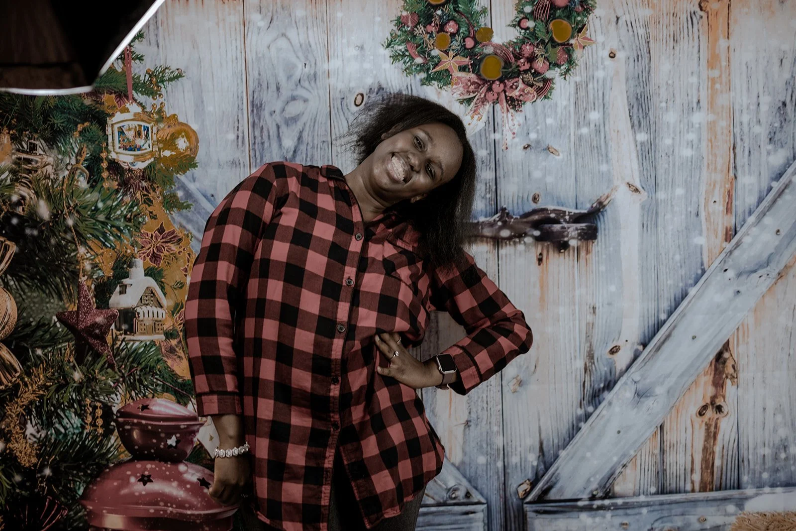 Woman in a red and black checkered shirt smiling, standing next to a decorated Christmas tree with ornaments and a festive background.