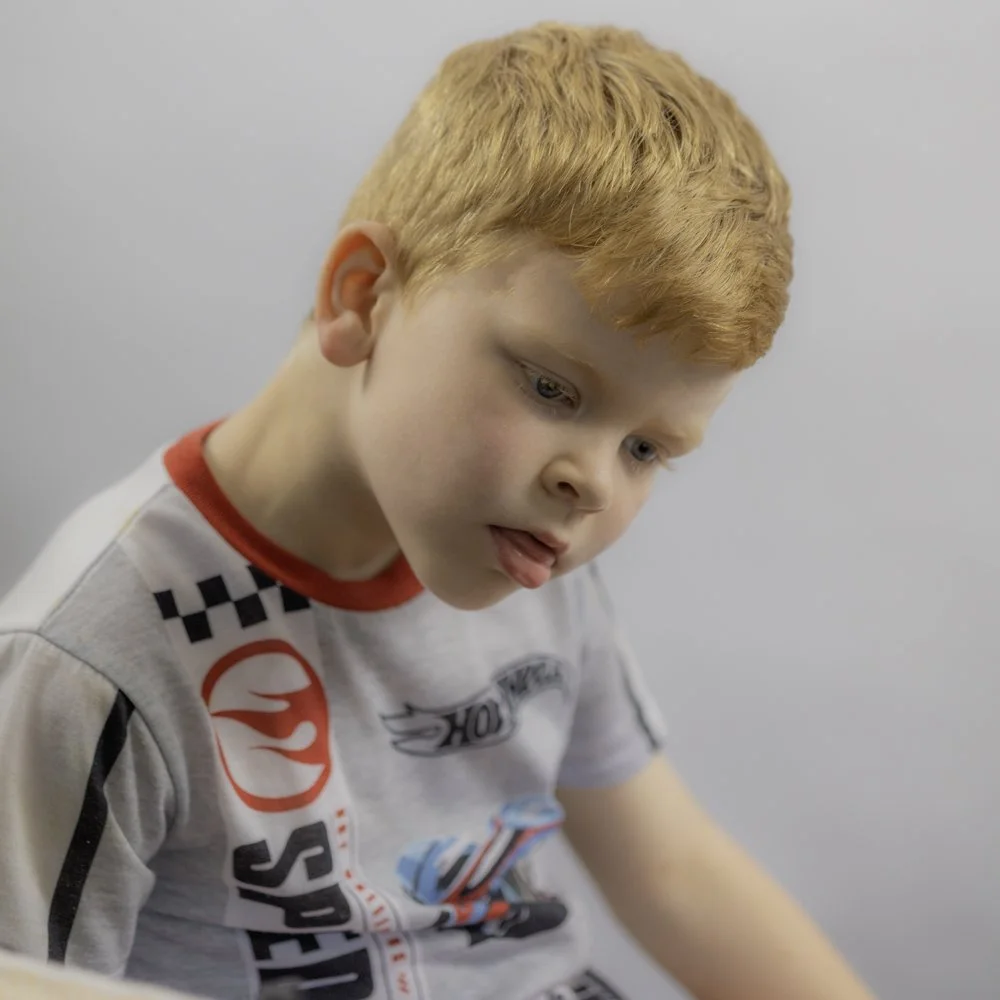 A young boy with red hair and light skin intently looking down, wearing a gray shirt with racing car and sponsor logos, against a plain gray background.