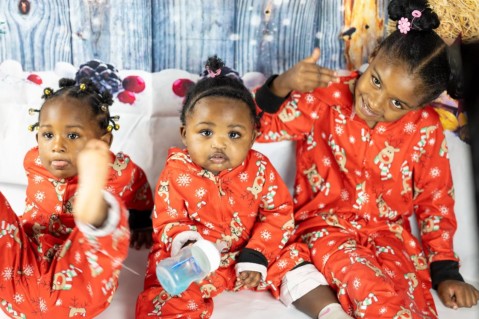 Three young girls sitting on a white bench, wearing Christmas-themed red pajamas with reindeer and snowflakes, during a holiday celebration. One girl is holding a baby bottle, all are making playful or curious expressions.