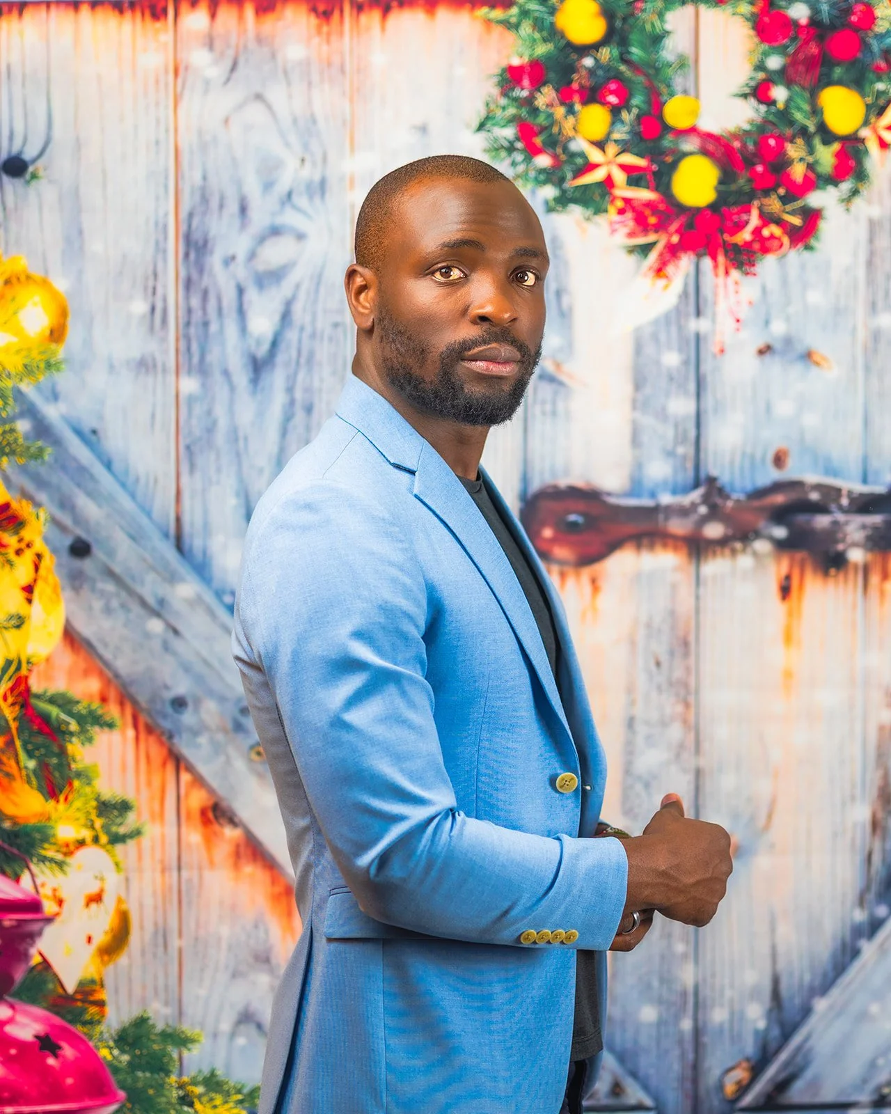 A man with a beard in a light blue blazer standing in front of a festive Christmas background with ornaments and decorations.