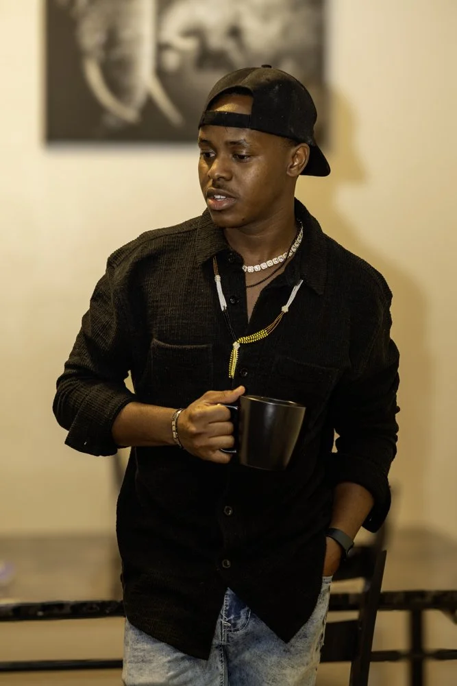 A young man wearing a black baseball cap backwards, a black button-up shirt, and light denim jeans, holding a black mug, standing indoors with a blurred background.