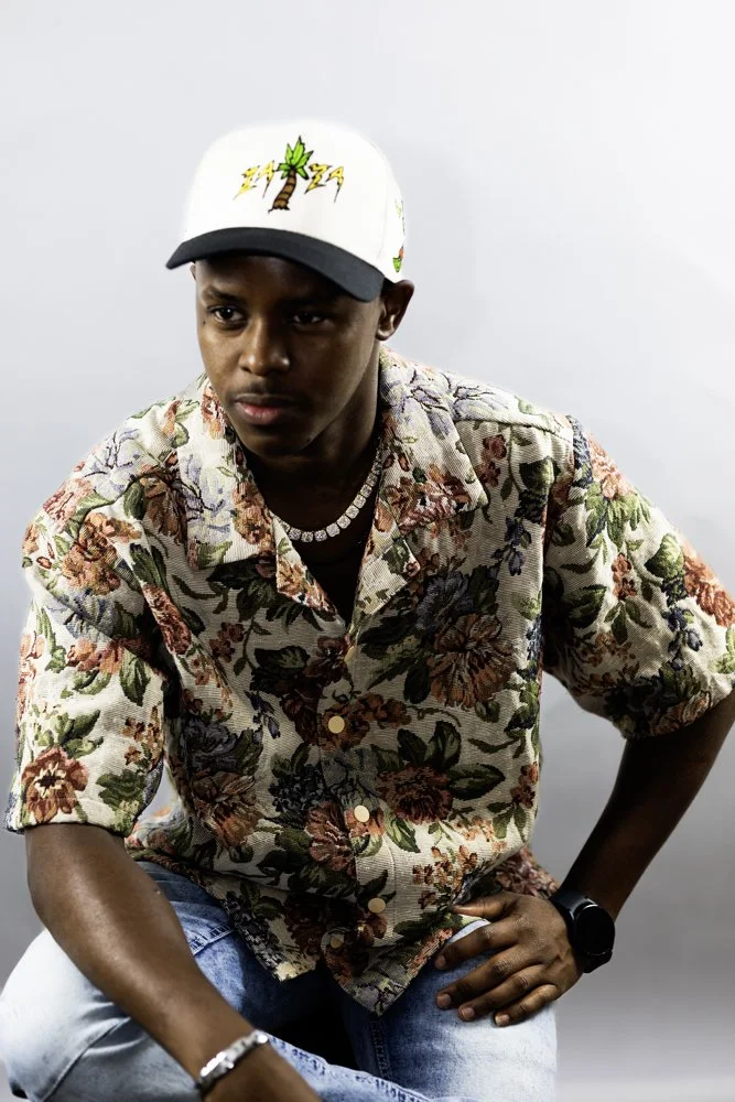 A young man wearing a floral patterned short-sleeve shirt, a white cap with a palm tree graphic, and accessories including a chain, watch, and bracelet, sitting against a plain background.